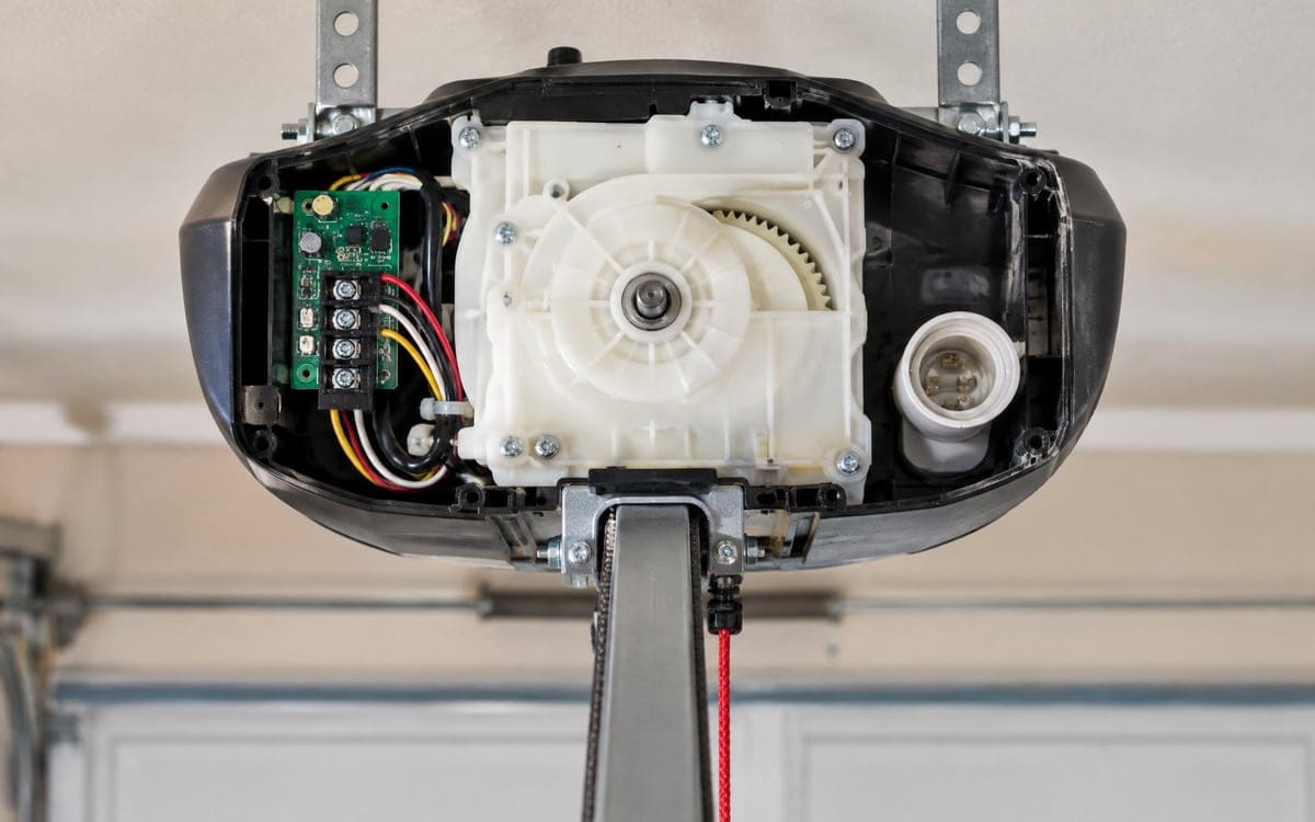An opener head with its cover off, showing the white drive gear housing and a green circuit board during garage door opener repair.