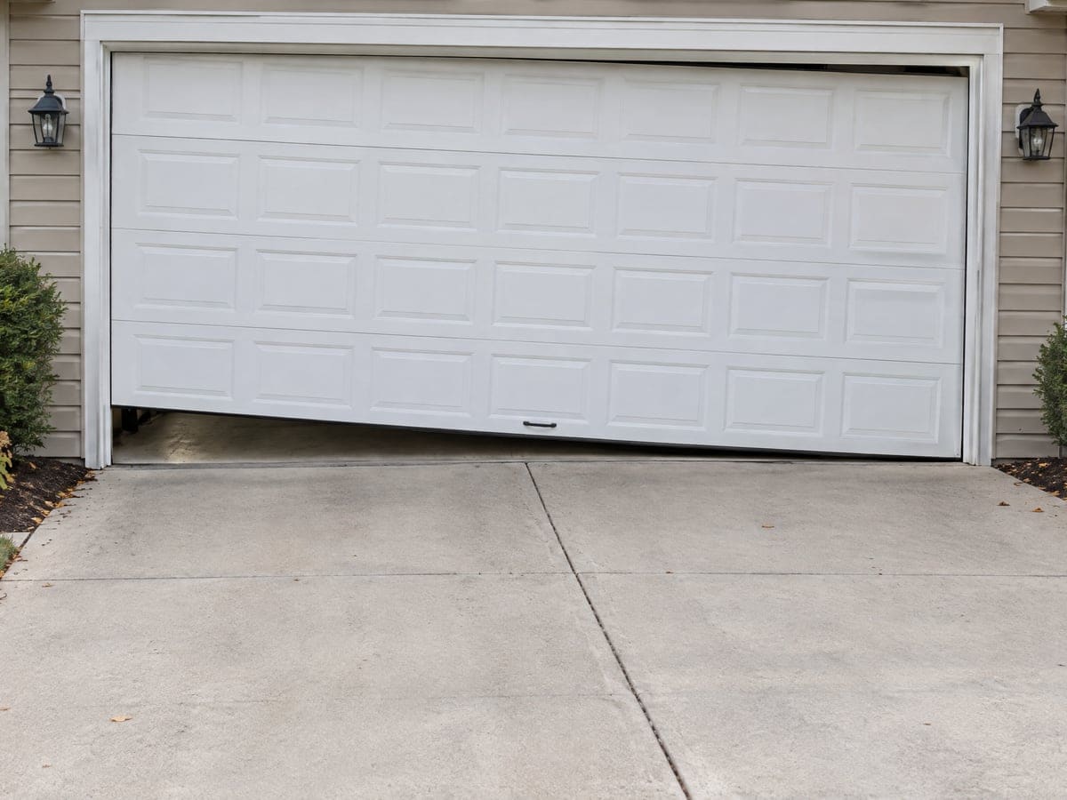 A garage door off track: a white two-car door hanging crooked in its frame, with a wedge-shaped gap under one end.