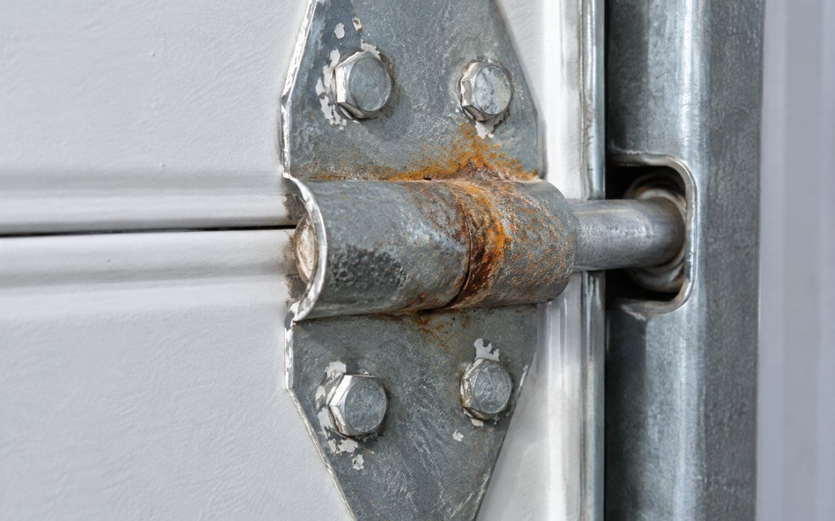 A rusted hinge pivot and roller stem on the inside of a steel door, one source of the squeaks behind a noisy garage door.